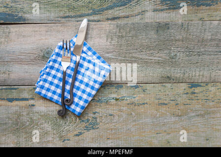 Forged handmade knife and fork and checkered blue-and-white napkin lie on a rough wooden background, with space for text Stock Photo