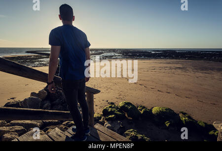 Young man standing on pier with Sanlucar de Barrameda beach in the background, looking to the horizon Stock Photo