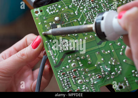 The girl-engineer repairs a switching power supply. Installation and soldering of electronic components using a soldering iron with a ceramic heater. Stock Photo