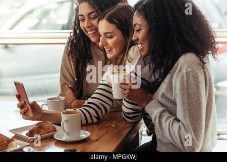 Three women sitting in a restaurant looking at mobile phone and smiling. Friends sitting at a cafe with coffee and snacks on the table looking at a mo Stock Photo