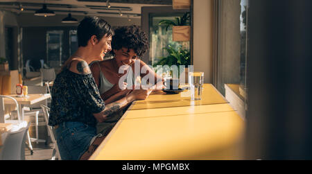 Two women sitting at cafe engrossed in using a smart phone. Female friends looking at mobile phone and smiling. Stock Photo