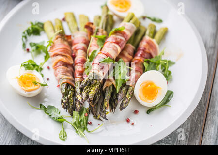 Fresh asparagus wrapped in bacon  on a white plate with arugula tomatoes and eggs. Stock Photo