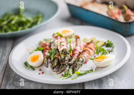 Fresh asparagus wrapped in bacon  on a white plate with arugula tomatoes and eggs. Stock Photo