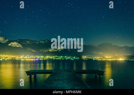 Nocturnal view of Lake Annecy, overlooked by mountains under stars of night sky, as seen by sitting on a wooden dock in the Sevrier region of Annecy. Stock Photo