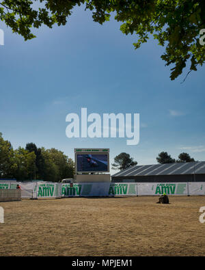 19-20th May 2018. Le Mans, France.  Behind the scenes at the MotoGP.  A young couple sit on the dirt ground to watch the practice sessions on one of the big screens.  The photograph is framed  with the leaves of an overhanging Oak tree. Stock Photo