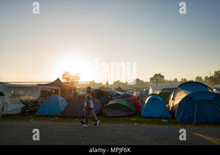 19-20th May 2018. Le Mans, France.  Behind the scenes at the MotoGP.  The sun sets over the race circuit and campsite in the late evening.  A couple walk past the campsite tents along the dirt road, and the perimeter of the race circuit can be seen in the distance on the horizon. Stock Photo