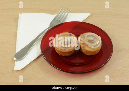 Quick breakfast of two small pinwheel cinnamon rolls on red glass plate with fork and folded napkin Stock Photo