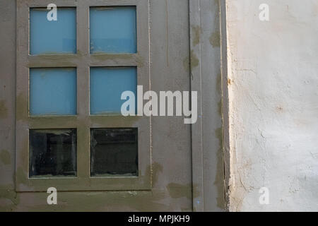 Dark gray metal door on the white plastered wall, in the door the squares of the glasses are blue and black. Stock Photo