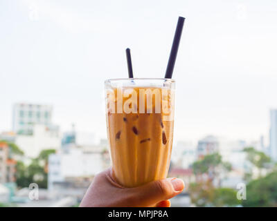 Ice Thai Milk Tea in Tall Glass with Dark Straw and Plastic Spoon in The Left Hand on Cityscape Background Stock Photo