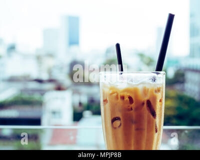 Ice Thai Milk Tea in Tall Glass with Dark Straw and Plastic Spoon inside in Glass Terrace on Cityscape Background Stock Photo