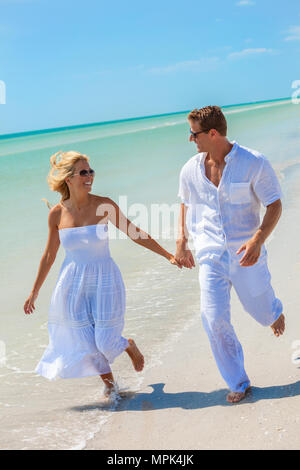 Man and woman romantic couple in white clothes holding hands running in the sea water on a deserted tropical beach with bright clear blue sky Stock Photo