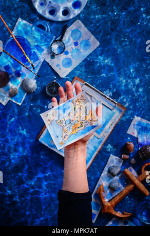 Artist holding a hand-painted fantasy island map. Marine watercolor concept. Creative artist workplace flat lay. Stock Photo