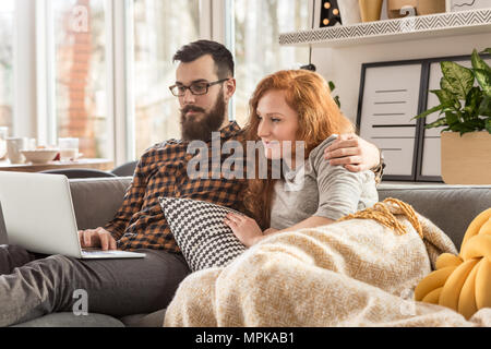 Man with laptop hugging his smiling wife lying under a blanket at home Stock Photo