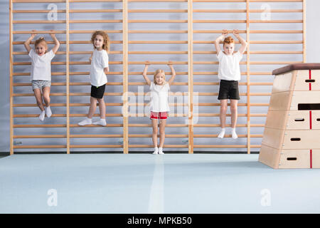 Young, active school boys and girls standing and hanging from wall bars, warming up for physical education athletics class Stock Photo