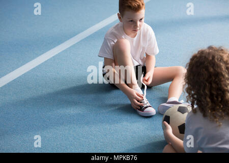 Portrait of a young boy sitting on a blue gym floor across from a friend holding a football and tying sport shoes Stock Photo