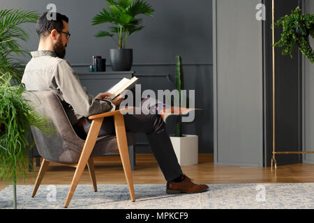 Man with beard sitting in grey armchair and reading a book in modern interior with plants Stock Photo