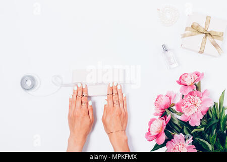 Woman packing gifts next to pink peony flowers, pearls and perfume bottle, top view. Female hands tying ribbon on present box, festive flat lay compos Stock Photo
