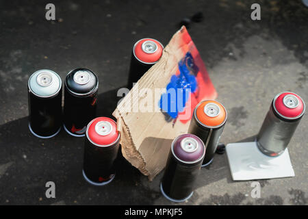 Aerosol metal cans of spray paint and cardboard palette for paint, graffiti on dark background of asphalt. Urban contemporary iconic culture of street youth Stock Photo