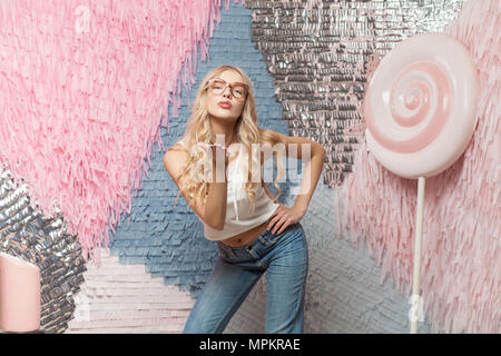 Beautiful young blonde girl with long hair in stylish red glasses on colorful abstract background with huge candy, dressed in a white top and jeans, s Stock Photo