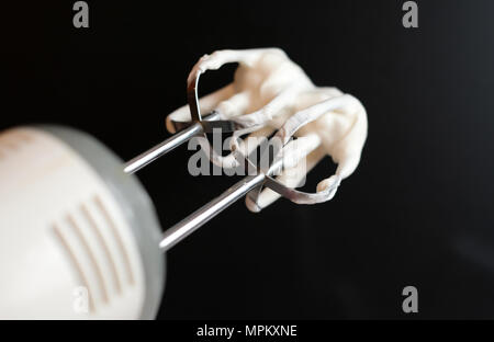 Electric hand mixer with whipped cream on the whisk or beater on a black background, selective focus. Dessert cooking or baking at home. Stock Photo