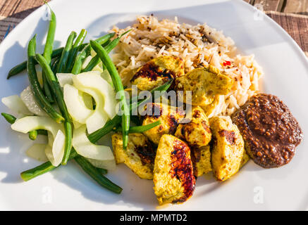 Indonesian meal served on white dinner plate on patio table in sunshine. Chicken satay, peanut sauce, kecap masin sauce, rice, green beans & cucumber Stock Photo