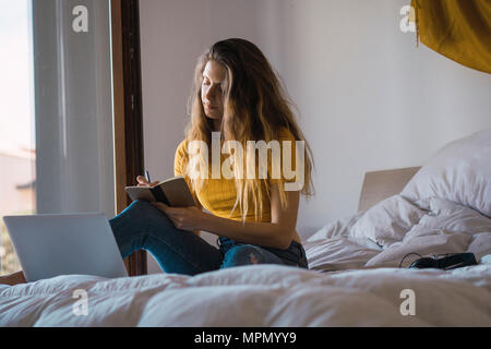 Young woman with laptop sitting on bed writing down something Stock Photo