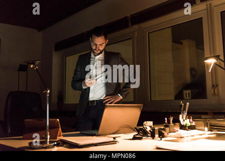Businessman standing in office at night using smartphone Stock Photo