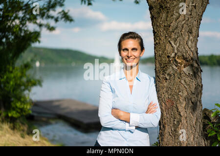 Portrait of smiling woman leaning against a tree at a lake Stock Photo