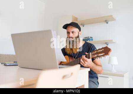 Serious young man at home with earphones and laptop playing guitar Stock Photo