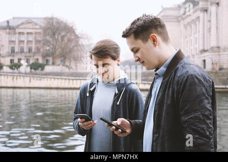 two antisocial mobile phone addicted male teenagers standing together using smartphone, technology concept, urban riverside location in Berlin Germany Stock Photo