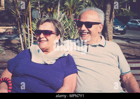 adults couple of man and woman sitting on a bench in urban area with parked cars and jardin on the background. both smiling and having a nice day with Stock Photo