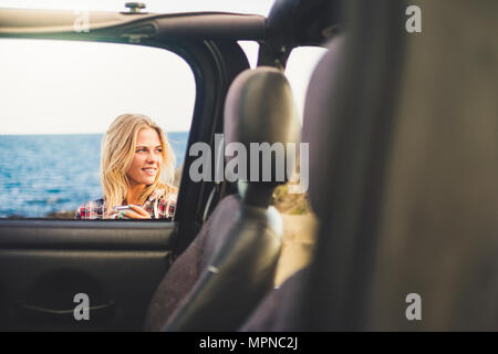 traveler blonde long hair girl take a mug with hot drink coffee or tea outside an open off road car. Ocean background and nice smile. Indipendent trav Stock Photo