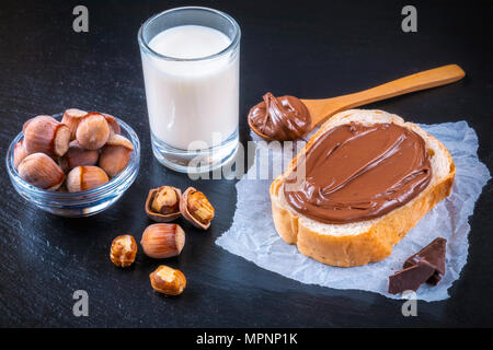 Slice of bread with spread chocolate cream and hazelnuts on black table. Serving a delicious breakfast concept. Focus on hazelnut in bowl. Stock Photo