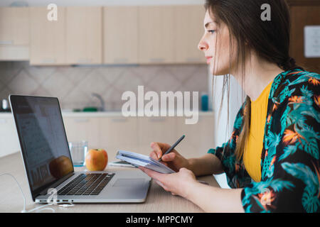 Young woman sitting at table at home using laptop and taking notes Stock Photo