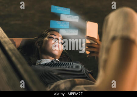 Young woman lying on bench at night with text emerging from smartphone Stock Photo