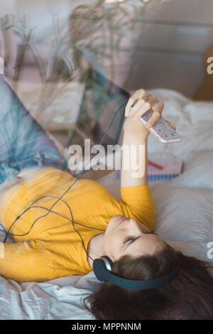 Young woman with headphones lying on bed taking selfie with smartphone Stock Photo