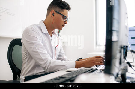 Software developer sitting at his office desk working on laptop wearing earphones. Man wearing spectacles working on laptop computer in office. Stock Photo