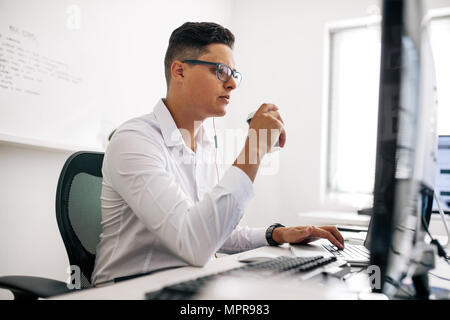 Smiling software developer sitting at his office desk working on laptop wearing earphones. Man wearing spectacles working on laptop computer in office Stock Photo