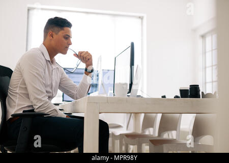 Software developer sitting at his office desk looking at laptop computer and thinking with spectacles in mouth. Man holding spectacles working on comp Stock Photo