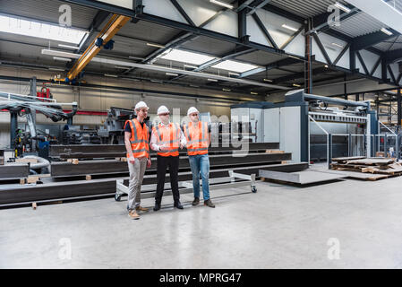Three men wearing hard hats and safety vests talking on factory shop floor Stock Photo