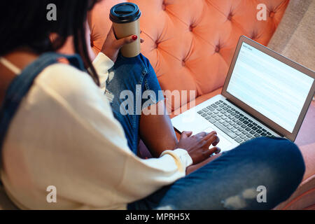 Young woman sitting on the couch with coffee to go using laptop Stock Photo