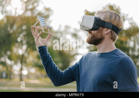 Man in autumnal park wearing Virtual Reality Glasses looking at miniature beach lounger Stock Photo