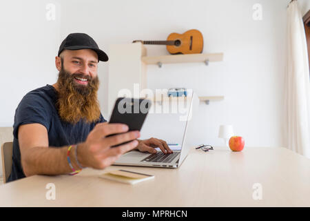 Portrait of bearded young man sitting at desk with laptop looking at cell phone Stock Photo