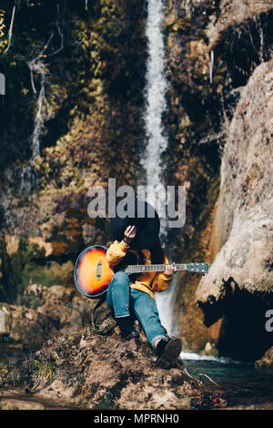 Spain, Lleida, young woman with guitar sitting on rock in front of waterfall covering face with hat Stock Photo