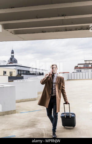 Businessman with rolling suitcase and smartphone walking at parking garage Stock Photo