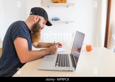 Bearded young man sitting at desk with laptop writing down something Stock Photo