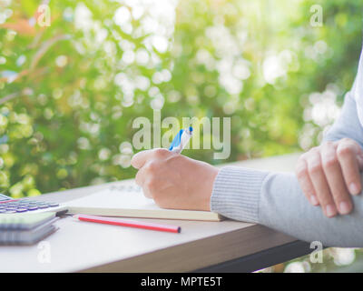 Woman hand writing with white pen, red pencil and calculator on working table and tree leaves background. Business and education concept. Stock Photo