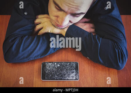 A man looks at his broken phone lying on a wooden table with a sad look. guy was upset about breaking his phone. Shattered phone display Stock Photo