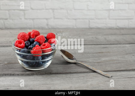 Small glass bowl full of blueberries with raspberries on top, with silver spoon next to it suggests its ready to eat, placed on gray wood desk. Stock Photo