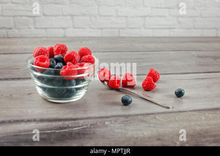 Silver spoon full of raspberries and blueberries, some spilled on gray wood desk, with out of focus bowl full of more berries on the side. Stock Photo